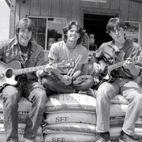 Black-and-white photo: three teenage musicians sit on stacked feed sacks in front of a rural feed store, two with guitars and one with drumsticks.