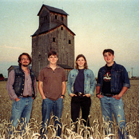 Faded color photo: four young musicians stand in a wheat field at dusk with a tall grain elevator behind them.