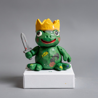 Lumpy green clay frog sitting upright with a crooked yellow crown and a tiny gray sword, uneven eyes and a red painted smile.
