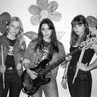 Black-and-white flash photo: three young women pose against a cinderblock wall painted with big flowers, one holding a bass guitar and one holding drumsticks.
