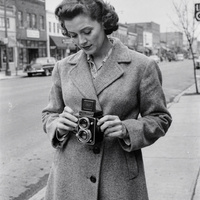Ruth Calder on Main Street, 1955. A black and white photograph from the 1950s of a woman in her thirties with short wavy hair and a wool coat, holding a twin-lens reflex camera at waist height on the sidewalk of a small-town main street, looking down into the camera's viewfinder.