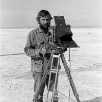 Sol Becker on the salt flats, 1976. A black and white photograph from the 1970s of a bearded man in his thirties in a canvas work jacket, standing behind a large-format wooden view camera on a tall tripod on a vast flat salt plain, holding the dark focusing cloth, a pale empty horizon behind him.