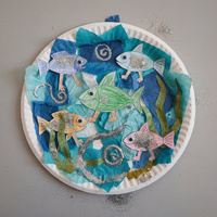 Round paper plate collage of an ocean in torn blue tissue paper, with paper fish with googly eyes around a big green fish, seaweed, and swirls of glitter glue.