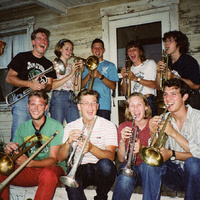 Color flash photo: about ten young people crowd onto the porch of an old wooden house with trombones and trumpets, laughing.